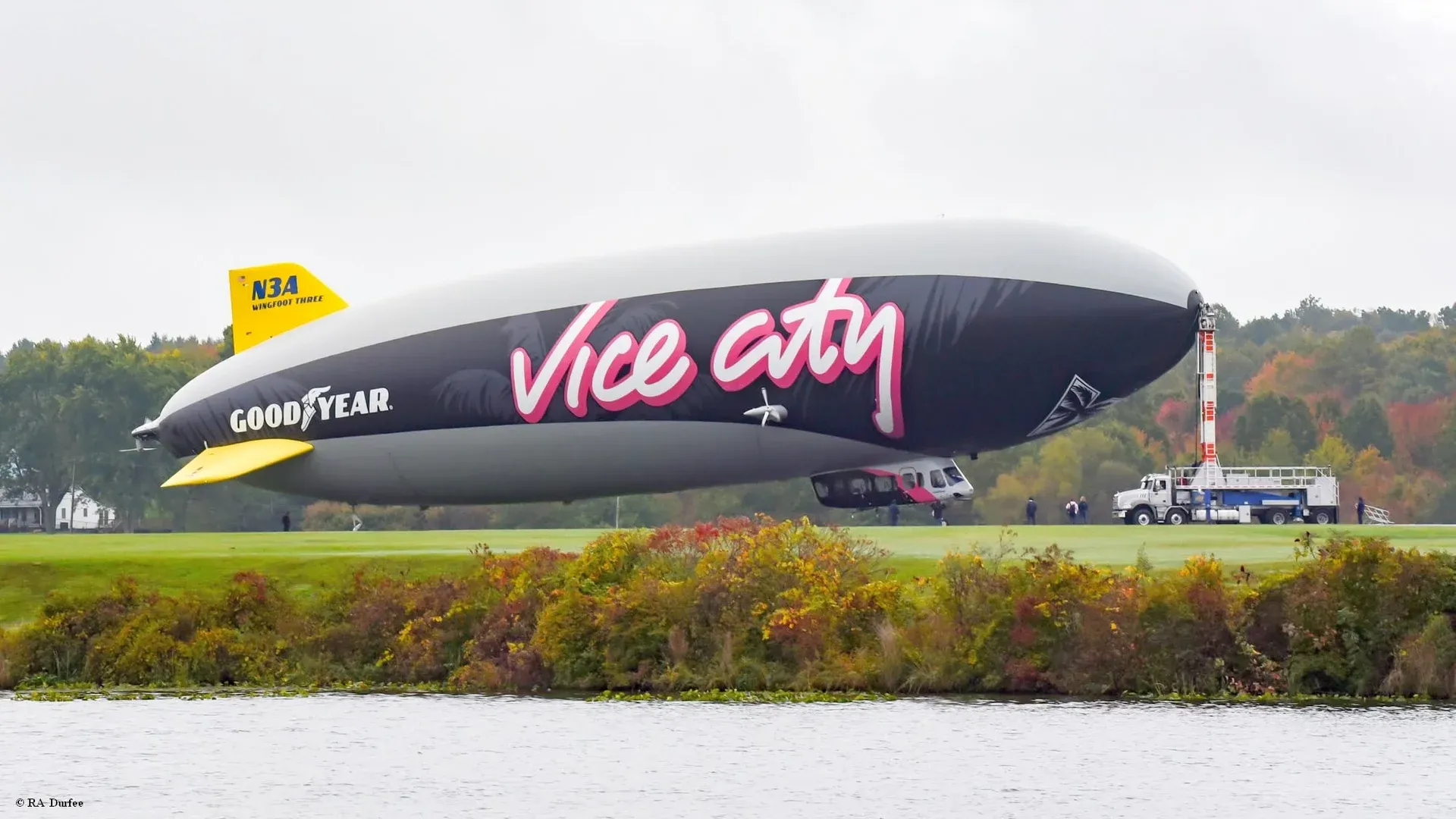 Photo by RA Durfee: Goodyear's airship Wingfoot Three, black with "Vice City" in white and pink script and Goodyear's name on its side, moored to its mast truck on a grass field beside a lake, with autumn trees behind